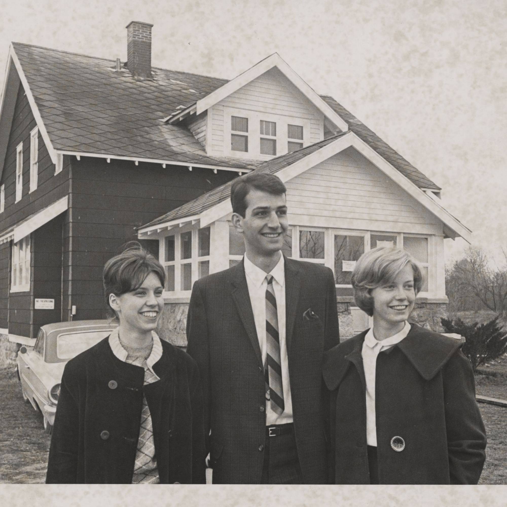 black and white photograph of young man standing between two women in front of home